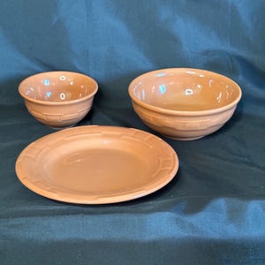 May include: A set of three terracotta-colored ceramic dishes. The set includes two bowls of different sizes and a plate. The bowls and plate have a textured, basket-weave design around the edges. The dishes are arranged on a dark green surface.