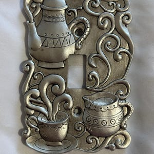 May include: Silver-colored light switch cover with a coffee and tea theme. The cover features a teapot, a cup of coffee with steam, and a creamer. Decorative swirls and patterns surround the central opening for the switch.