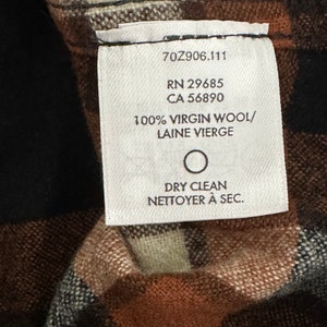 May include: A white tag with black text on a brown and black plaid fabric. The tag reads "70Z906.111 RN 29685 CA 56890 100% VIRGIN WOOL/ LAINE VIERGE DRY CLEAN NETTOYER &Agrave; SEC."