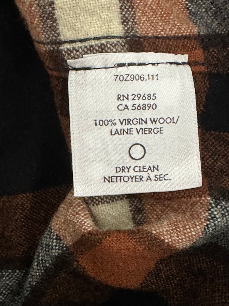 May include: A white tag with black text on a brown and black plaid fabric. The tag reads "70Z906.111 RN 29685 CA 56890 100% VIRGIN WOOL/ LAINE VIERGE DRY CLEAN NETTOYER &Agrave; SEC."