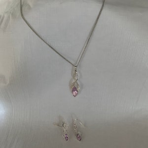 May include: A silver necklace and earrings set with a purple gemstone pendant and matching earrings. The pendant and earrings feature a delicate, swirling design.