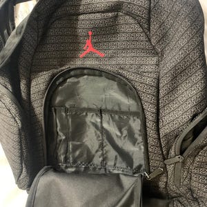 K&ouml;nnte beinhalten: Schwarzer Rucksack mit einem roten gestickten Logo eines Basketballspielers. Der Rucksack hat mehrere Taschen und Rei&szlig;verschl&uuml;sse. Der Stoff hat ein sich wiederholendes Muster der Zahl "23", die in Wei&szlig; gedruckt ist.
