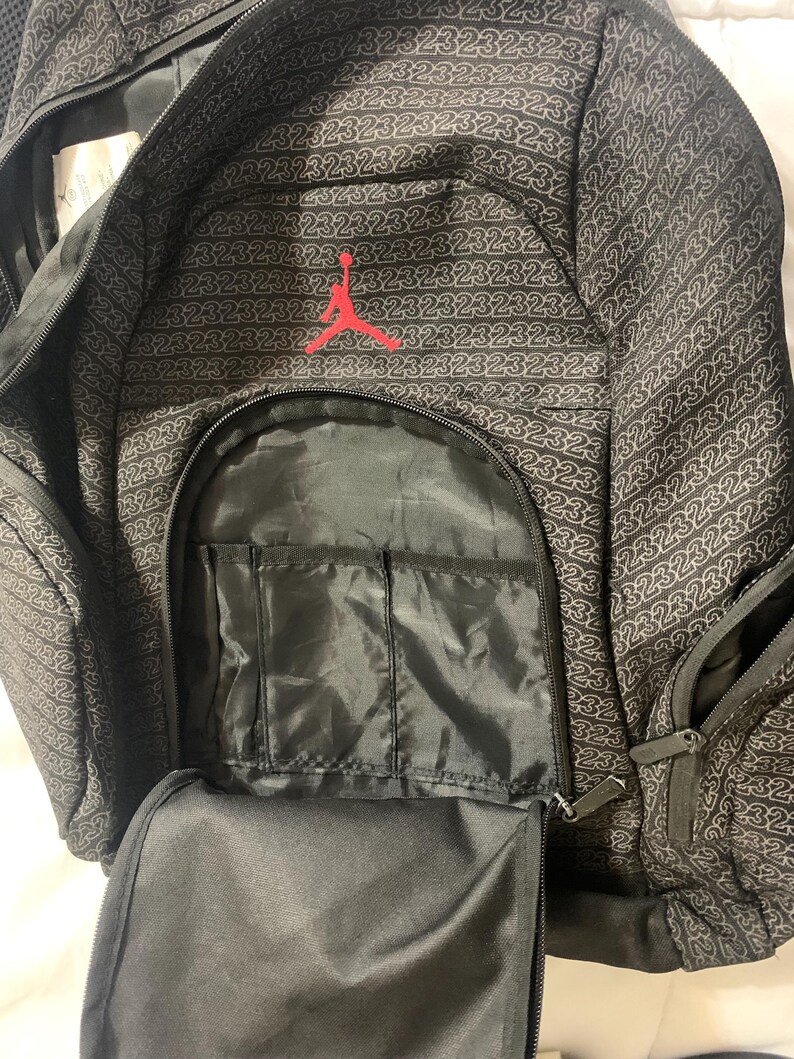 K&ouml;nnte beinhalten: Schwarzer Rucksack mit einem roten gestickten Logo eines Basketballspielers. Der Rucksack hat mehrere Taschen und Rei&szlig;verschl&uuml;sse. Der Stoff hat ein sich wiederholendes Muster der Zahl "23", die in Wei&szlig; gedruckt ist.