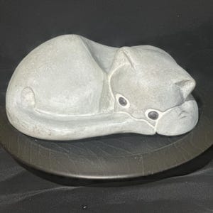 May include: A light gray stone sculpture of a cat in a curled, sleeping position. The figure features smooth, rounded contours, pointed ears, and two small, dark, circular inlaid eyes. The cat is depicted resting with its tail tucked along its side.