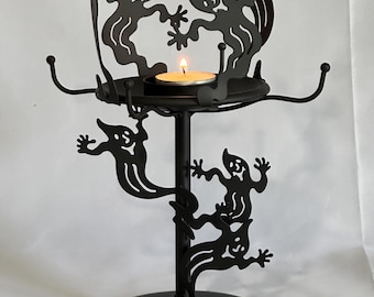 May include: Black metal Halloween candle holder featuring whimsical ghost silhouettes. The candle holder has a round base, a central stem, and a circular platform to hold a lit candle. The design includes decorative hooks.