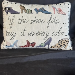 May include: A rectangular woven throw pillow with a cream background and a dark border. The pillow features illustrations of eight different women's shoes in various colors and patterns, with the text "If the shoe fits... buy it in every color..."