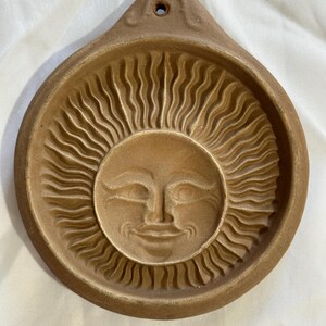 May include: A brown ceramic sun-shaped wall hanging. The sun has a smiling face in the center, surrounded by wavy rays. The top has a small hole for hanging. The piece is a warm, earthy tone.