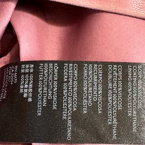 Op de afbeelding: Een close-up van een zwart kledinglabel met witte tekst. Het label geeft de samenstelling van de stof van een kledingstuk weer, waaronder viscose, polyurethaan en polyester. De achtergrond is een roze stof.