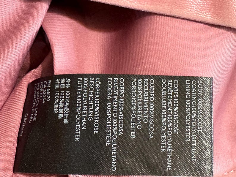 Op de afbeelding: Een close-up van een zwart kledinglabel met witte tekst. Het label geeft de samenstelling van de stof van een kledingstuk weer, waaronder viscose, polyurethaan en polyester. De achtergrond is een roze stof.