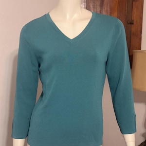May include: Teal-colored V-neck sweater with three-quarter sleeves. The sweater has a ribbed texture on the front and a button detail at the cuffs. The garment is displayed on a mannequin.