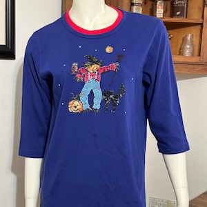 May include: A blue, long-sleeved t-shirt with a festive Halloween design featuring a scarecrow, an owl, a black cat, and a pumpkin. The design is made of sequins and rhinestones.