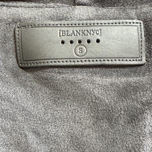 Pu&ograve; includere: Primo piano di un tessuto grigio simile alla pelle scamosciata con un'etichetta rettangolare cucita. L'etichetta recita "[BLANKNYC]" sopra tre piccoli punti e la lettera "S" in un cerchio. Il tessuto ha un aspetto morbido e strutturato.