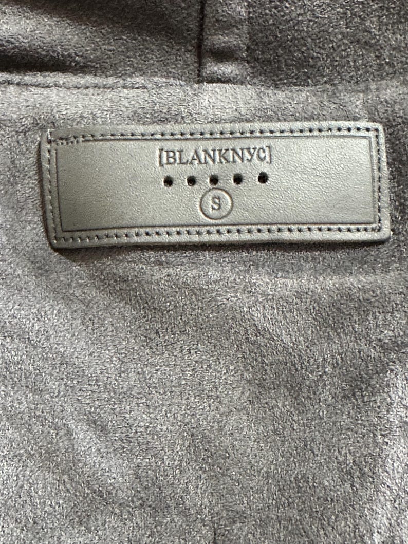 Pu&ograve; includere: Primo piano di un tessuto grigio simile alla pelle scamosciata con un'etichetta rettangolare cucita. L'etichetta recita "[BLANKNYC]" sopra tre piccoli punti e la lettera "S" in un cerchio. Il tessuto ha un aspetto morbido e strutturato.