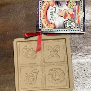 May include: A brown ceramic cookie mold with four different Christmas designs: an ornament, candy canes, a horn, and a wreath. The mold is on a wooden surface with a red ribbon tied around it. The text on the packaging reads "Brown Bag Cookie Art Cookie & Craft Mold".