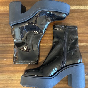 May include: A pair of black patent leather ankle boots with a chunky platform heel and a zipper on the back. The boots are on a wooden surface.
