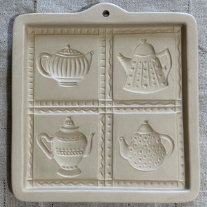 May include: A beige ceramic cookie press with four different teapot designs. Each teapot has a different pattern: one has vertical lines, one has hearts, one has a rope design, and one has dots.