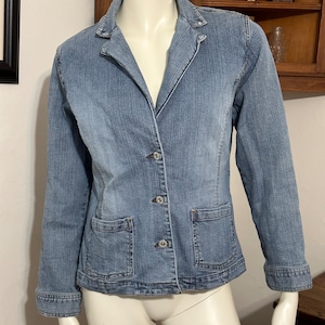 May include: A light blue denim blazer with a classic collar, three buttons, and two front pockets. The jacket is displayed on a mannequin. The denim fabric has a faded appearance.