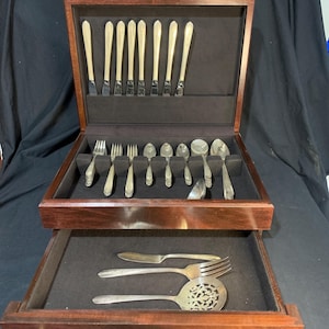 May include: A wooden silverware chest with two drawers, open to reveal a complete set of silver utensils. The top drawer holds knives, forks, and spoons, while the bottom drawer contains serving pieces. The chest is made of dark wood.