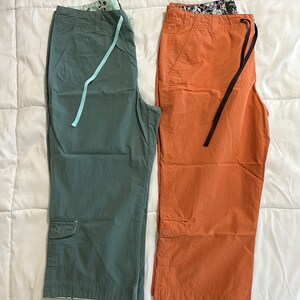 May include: Two pairs of cropped pants. One pair is a dark green color and the other pair is a burnt orange color. Both pairs have a cargo pocket on the left leg and a drawstring at the waist.