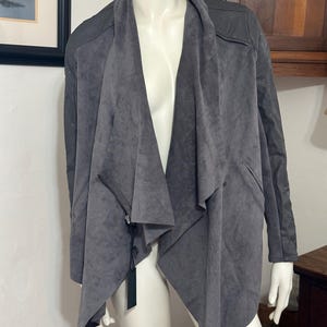 Pu&ograve; includere: Giacca grigia in similpelle scamosciata con parte anteriore drappeggiata e chiusura con zip. La giacca ha un collo a cascata e maniche lunghe. La giacca &egrave; esposta su un manichino. Un'etichetta &egrave; attaccata.
