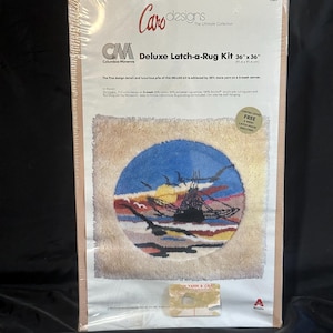 May include: A Caro Designs Deluxe Latch-a-Rug Kit measuring 36 inches by 36 inches. The packaging features a circular latch hook rug design depicting a ship at sea during sunset with birds flying overhead, all set against a cream-colored background.
