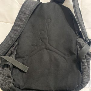 K&ouml;nnte beinhalten: Schwarzer Rucksack mit einem gro&szlig;en gestickten Jordan-Logo auf der Vorderseite. Der Rucksack hat ein Rei&szlig;verschluss-Hauptfach und ein kleineres Fach vorne. Der Stoff hat ein dezentes Muster mit der Nummer 23.