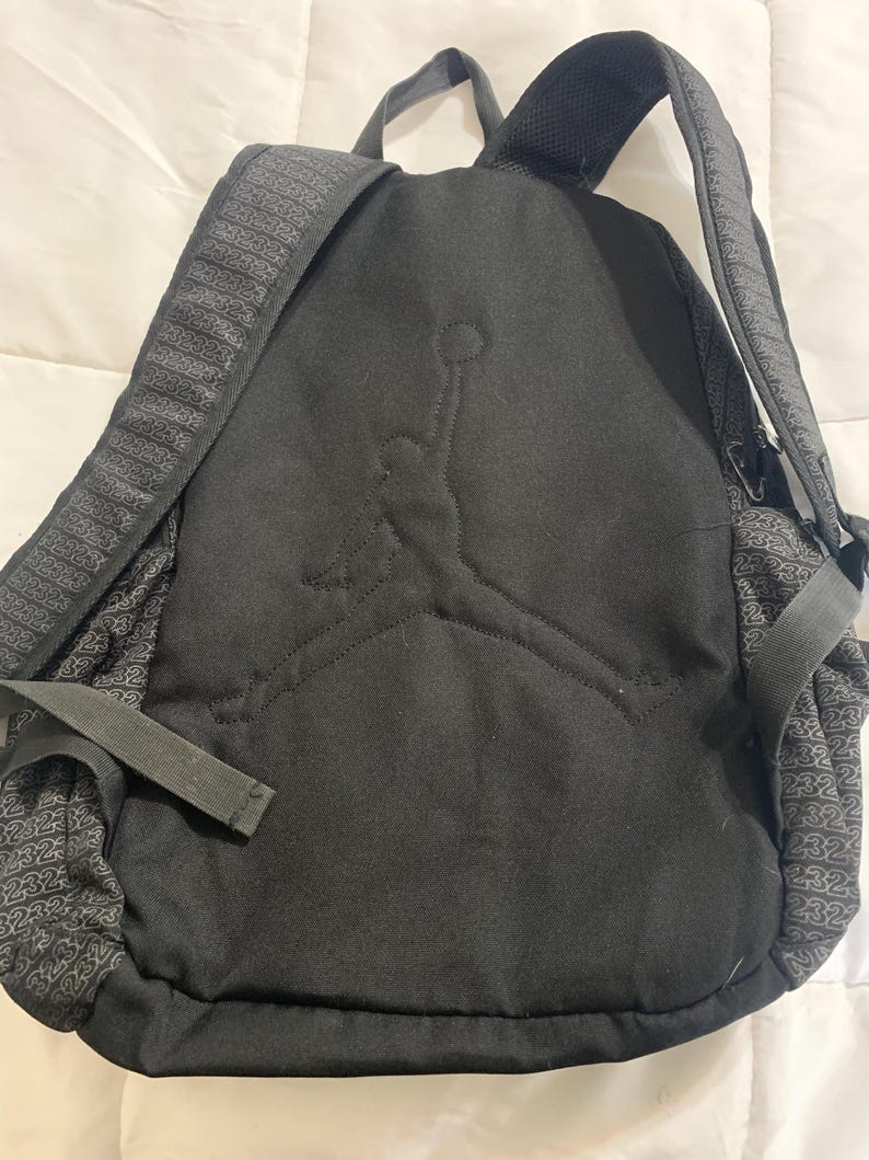 K&ouml;nnte beinhalten: Schwarzer Rucksack mit einem gro&szlig;en gestickten Jordan-Logo auf der Vorderseite. Der Rucksack hat ein Rei&szlig;verschluss-Hauptfach und ein kleineres Fach vorne. Der Stoff hat ein dezentes Muster mit der Nummer 23.
