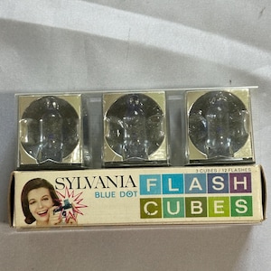 May include: Vintage Sylvania Blue Dot Flash Cubes in original packaging. The box features a retro image of a woman holding a camera. The box reads "FLASH CUBES" in colorful block letters. The package contains three flash cubes.