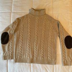 May include: A beige cable knit turtleneck sweater with brown leather elbow patches.