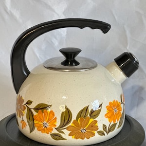 May include: Cream-colored vintage tea kettle with a black handle, spout, and lid. The kettle is decorated with orange and brown floral designs and sits on a dark gray tray. A classic kitchenware item.