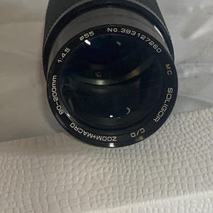 May include: Black Soligor zoom macro camera lens. The lens has the following specifications: 60-200mm, f/4.5, and a 55mm filter thread. The lens has the text "ZOOM+MACRO" and "MC SOLIGOR" printed on it.
