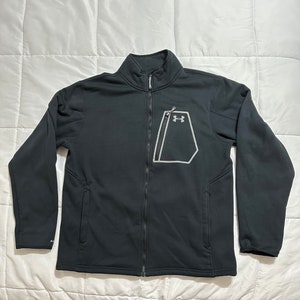 May include: Black zippered jacket with a gray Under Armour logo on the front left chest. The jacket has a stand-up collar and a zippered pocket on the left side.