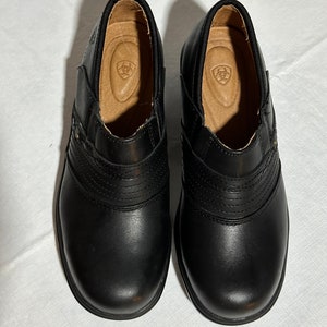 May include: A pair of black leather slip-on shoes. The shoes have a rounded toe, a stitched design across the top, and a small gold-colored stud on the side. The interior features a tan insole with a logo.