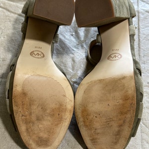 Pu&ograve; includere: Un paio di sandali verdi in camoscio con tacco e cinturini alla caviglia. Le suole sono in pelle marrone con il testo "8 1/2 M" e "MICHAEL KORS" stampato su di esse.
