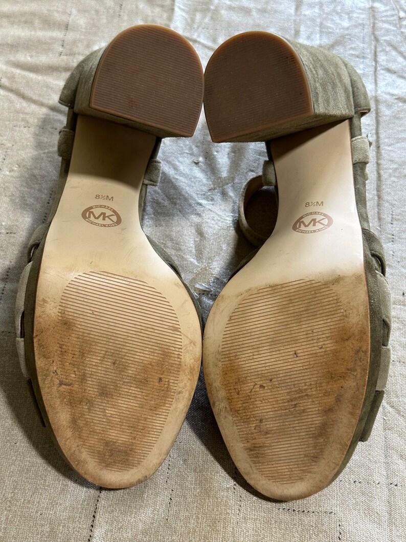 Pu&ograve; includere: Un paio di sandali verdi in camoscio con tacco e cinturini alla caviglia. Le suole sono in pelle marrone con il testo "8 1/2 M" e "MICHAEL KORS" stampato su di esse.