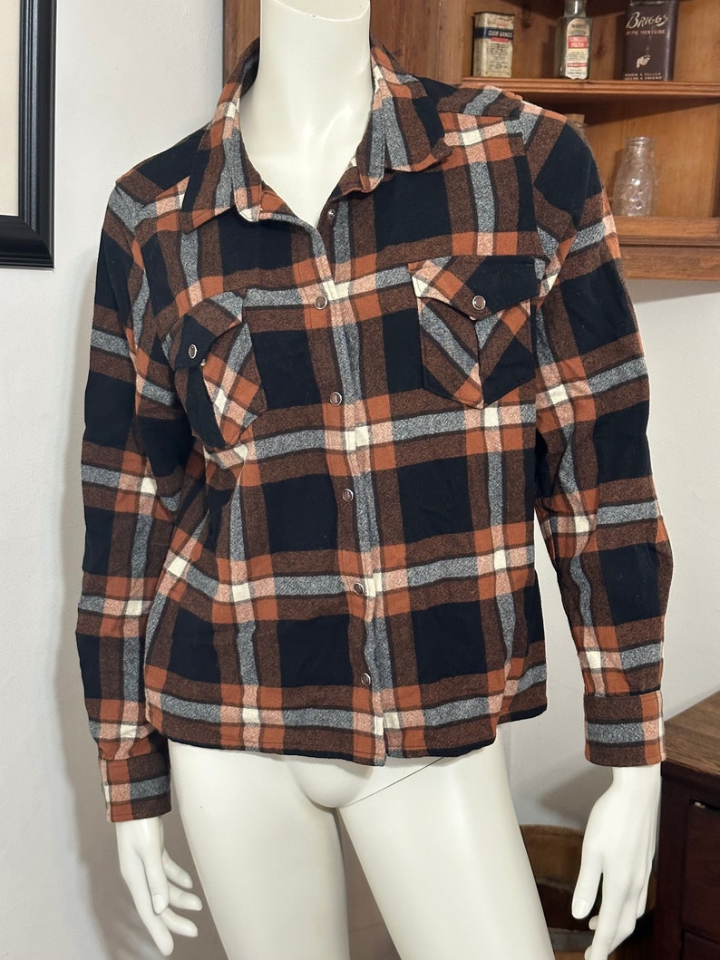 May include: A long-sleeved plaid shirt with a brown, black, and white checkered pattern. The shirt has a button-down front and two chest pockets.