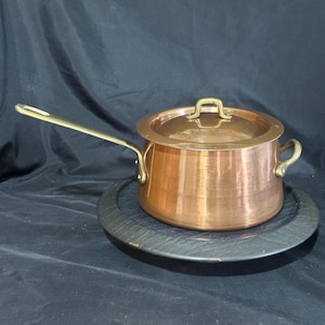 May include: A polished copper saucepan with a matching lid, featuring a long brass handle and a smaller brass side handle. The pot has a cylindrical body with a smooth, reflective surface and a circular lid with a central brass handle.