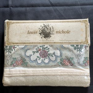 May include: A packaged twin flat sheet by Louis Nichole. The sheet features a floral pattern in shades of pink, green, and blue, with a cream-colored lace trim. The packaging includes the text "louis nichole" and "One Twin Flat Sheet."