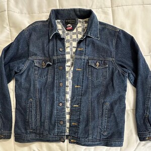 May include: A dark blue denim jacket with a button-up front, two chest pockets, and two side pockets. The jacket has a collared neckline and a plaid patterned lining. The brand name "OKCH" is visible on the collar.