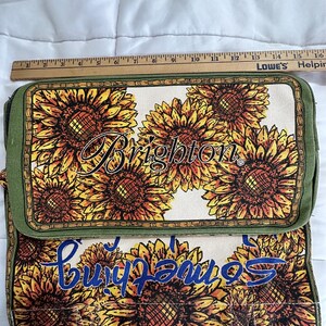 Peut inclure: Sac rectangulaire avec un motif de tournesols. Le sac pr&eacute;sente un fond cr&egrave;me avec de grands tournesols dans les tons de jaune, orange et marron. Le mot "Brighton" est &eacute;crit en cursive. Le sac mesure environ 30 cm de long.