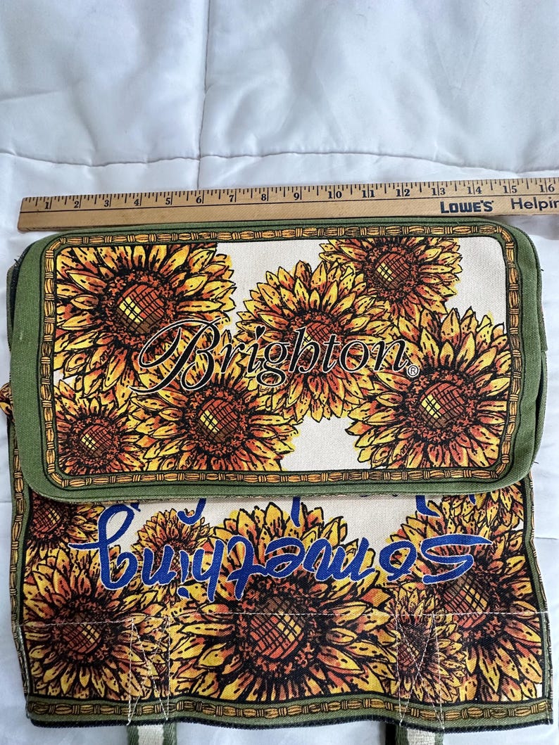 Peut inclure: Sac rectangulaire avec un motif de tournesols. Le sac pr&eacute;sente un fond cr&egrave;me avec de grands tournesols dans les tons de jaune, orange et marron. Le mot "Brighton" est &eacute;crit en cursive. Le sac mesure environ 30 cm de long.