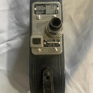 May include: Vintage Keystone Model B-1 16mm movie camera. The camera is dark blue with a silver-colored metal faceplate. The faceplate has dials, a lens, and the text "MODEL B-1" and "MADE IN U.S.A.". A black leather strap is attached.