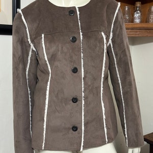 May include: A brown suede jacket with white trim around the edges. The jacket has a button closure and a fitted silhouette.