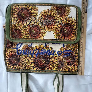 Peut inclure: Un sac fourre-tout rectangulaire avec un rabat, orn&eacute; d'un motif de tournesols sur un fond cr&egrave;me. Le sac a une bordure et des poign&eacute;es vertes. Le mot "Brighton" est imprim&eacute; sur le rabat, et les mots "Something" sont imprim&eacute;s sur le corps du sac.