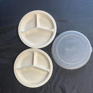 May include: Two round, light beige plastic plates, each featuring three divided sections for food separation. Beside the plates is a single, circular, translucent plastic lid with embossed text, designed to fit over the plates for storage.