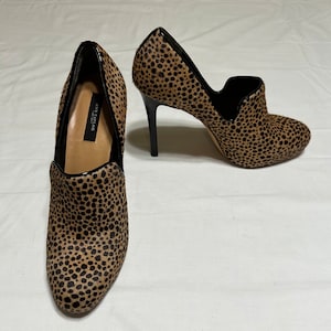 May include: A pair of brown and black leopard print heeled ankle boots with a black heel and a black trim around the ankle opening. The boots are made by the brand "Anne Klein" and have a pointed toe.