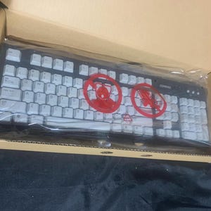 May include: A black computer keyboard with white keys, enclosed in a clear plastic protective sleeve inside a cardboard box. The keyboard features two prominent red circular warning symbols printed on the plastic film covering the keys.