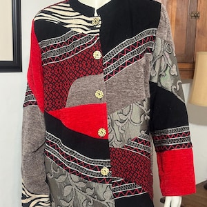May include: A long-sleeve jacket featuring a patchwork design of black, red, and gray fabric panels. The garment includes sections with floral patterns, geometric shapes, and zebra stripes, secured by five light-colored circular buttons.