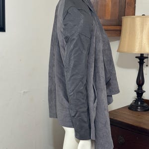 Pu&ograve; includere: Giacca grigia con design drappeggiato. La giacca ha un colletto e maniche lunghe. Il tessuto sembra avere una consistenza simile alla pelle scamosciata. La giacca &egrave; esposta su un manichino.