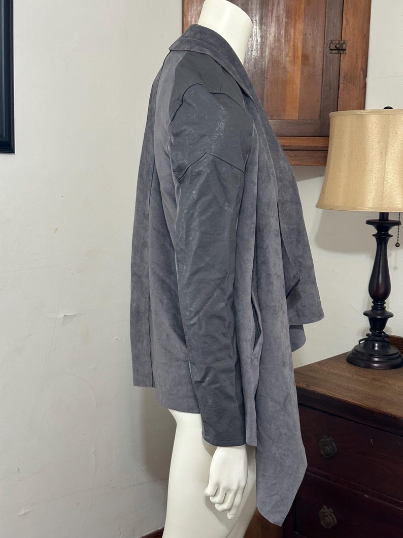 Pu&ograve; includere: Giacca grigia con design drappeggiato. La giacca ha un colletto e maniche lunghe. Il tessuto sembra avere una consistenza simile alla pelle scamosciata. La giacca &egrave; esposta su un manichino.